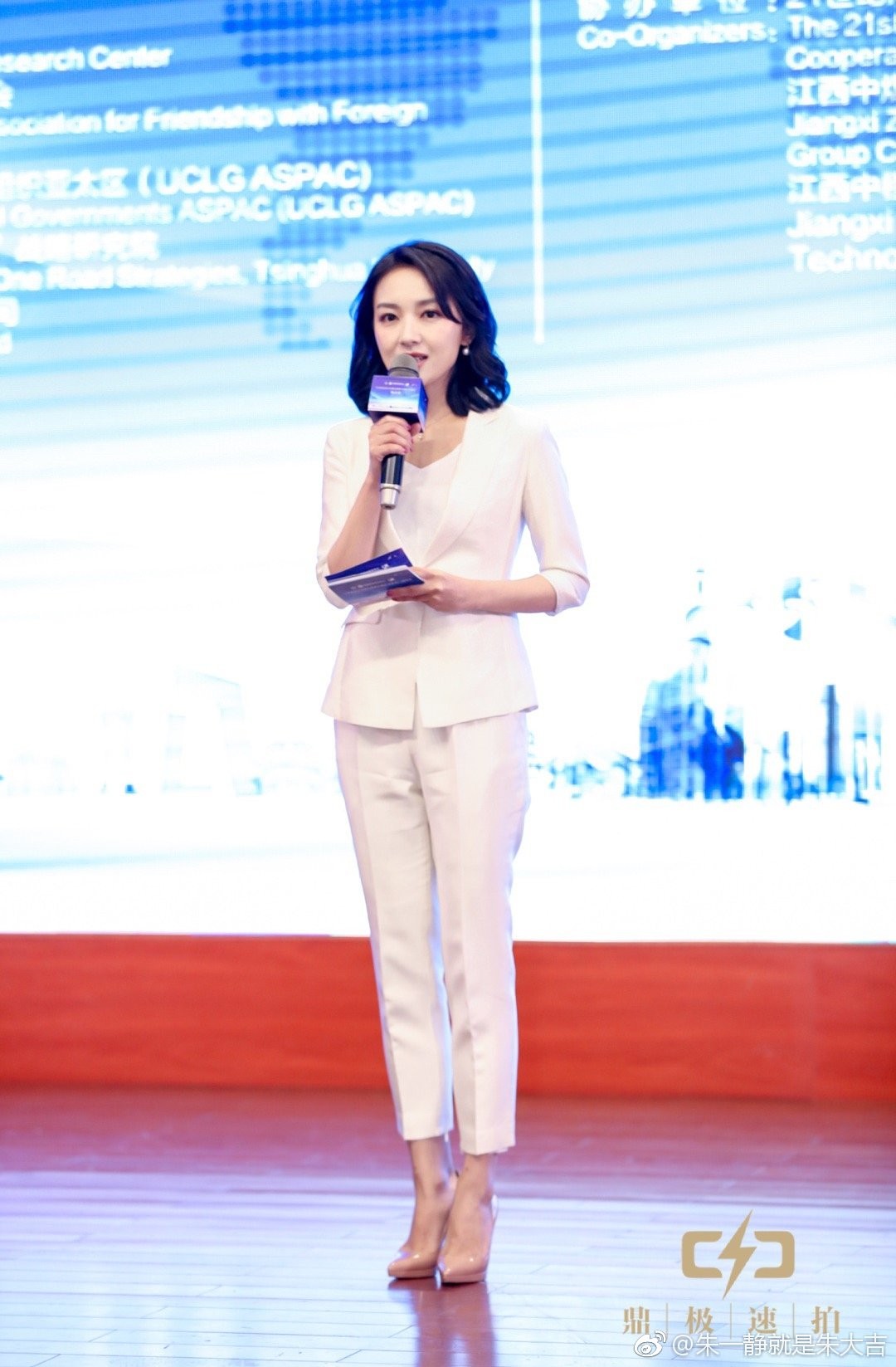 朱一静微笑注视镜头，身后舞台灯光璀璨，尽显主持人的沉稳与光彩