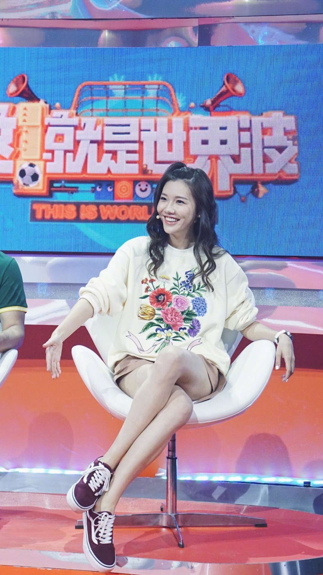 她手持话筒微笑交流，短裙与高跟鞋衬出修长身姿，眼神专注有光