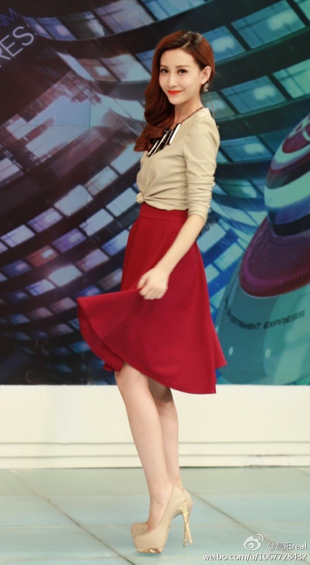 晨阳侧身立于演播台前，黑色职业装搭配丝袜与高跟鞋，手持话筒专注聆听