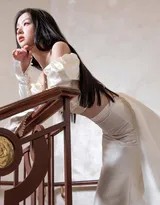 包上恩身着白色抹胸长裙，在楼梯转角处轻盈回眸，裙摆如云朵般铺开，光影洒落肩头