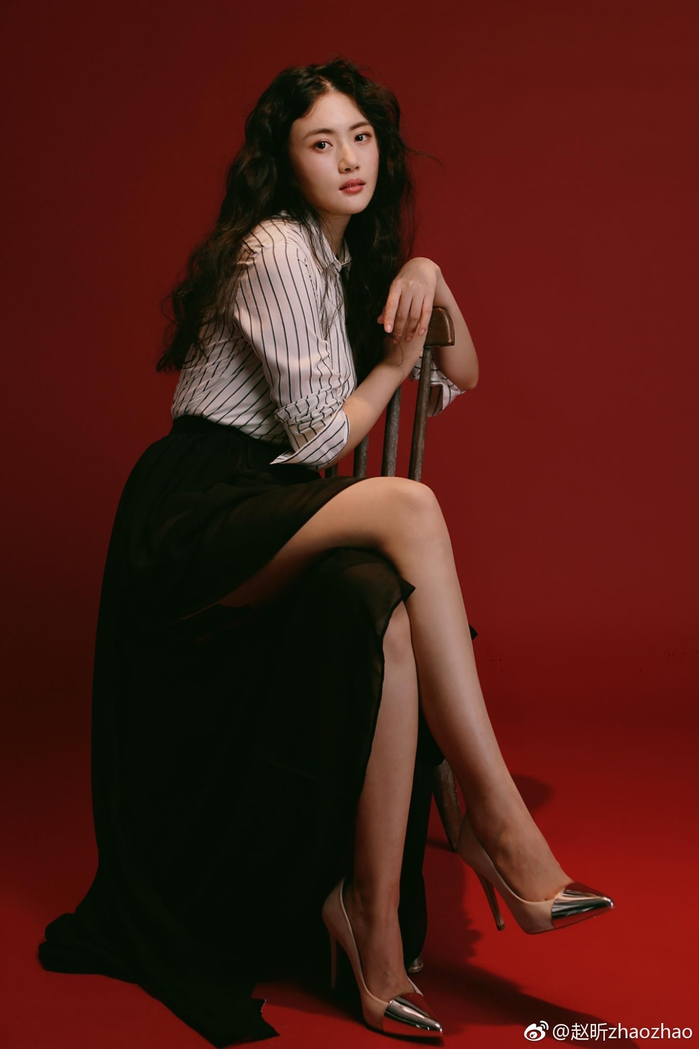 赵昕倚靠墙边，黑色皮裤在光影下反射出柔和光泽，双腿交叉更显线条修长，整体造型简约而富有冲击力